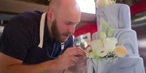 A baker in an apron carefully placing white flowers onto a grey marbled wedding cake.