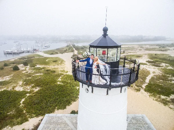 A bride and groom posing on the observation deck of a lighthouse overlooking a harbor.