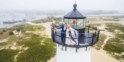 A bride and groom posing on the observation deck of a lighthouse overlooking a harbor.