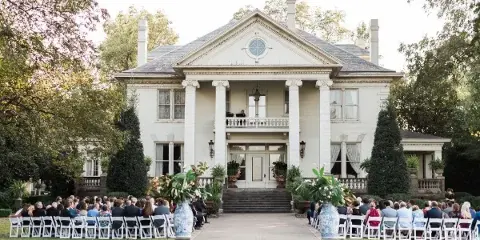Exterior view of a white Greek Revival mansion with a wedding ceremony setup in the front yard.
