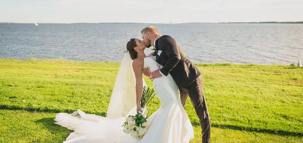 A bride and groom kissing on a grassy lawn with a large body of water in the background.