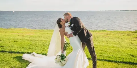 A bride and groom kissing on a grassy lawn with a large body of water in the background.