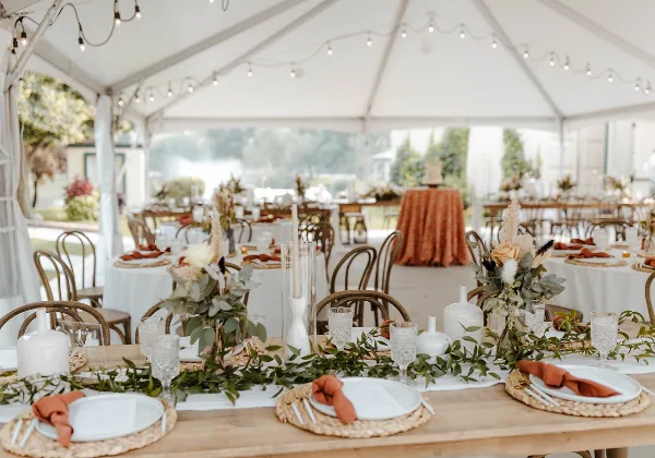 A wedding table setting under a white tent featuring white plates, glassware, and floral centerpieces.
