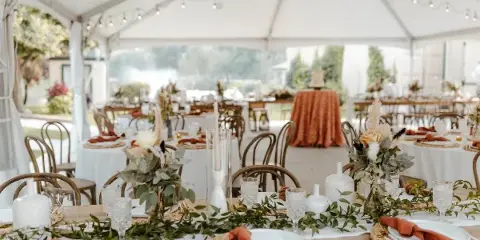 A wedding table setting under a white tent featuring white plates, glassware, and floral centerpieces.