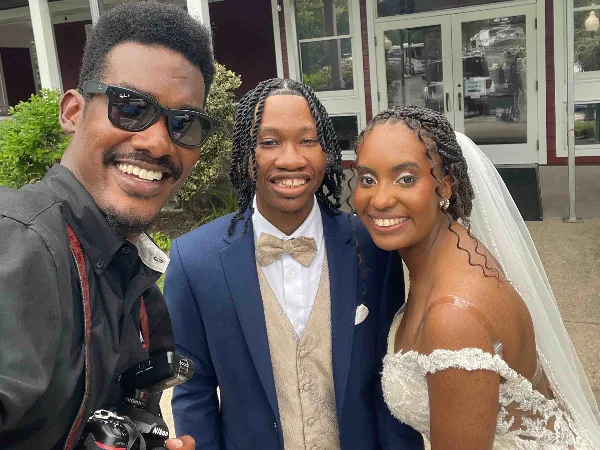 A wedding photographer holding a camera takes a selfie with a bride and groom outside a venue.