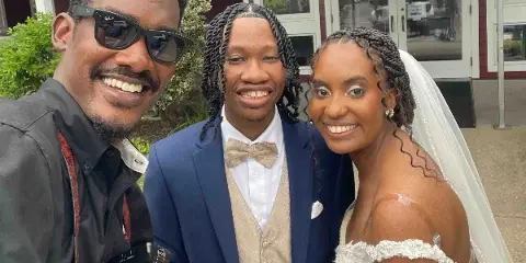 A wedding photographer holding a camera takes a selfie with a bride and groom outside a venue.