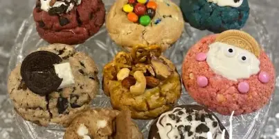 A glass stand holding an assortment of decorated cookies, including M&M cookies, Oreo cookies, and pretzel nut cookies, with a heart-shaped frame in the background.