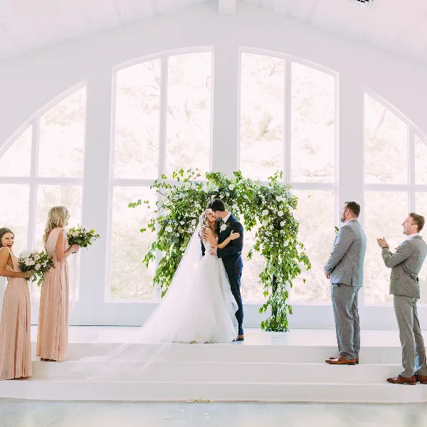 A bride and groom share a kiss at the altar during a wedding ceremony, surrounded by bridesmaids and groomsmen.
