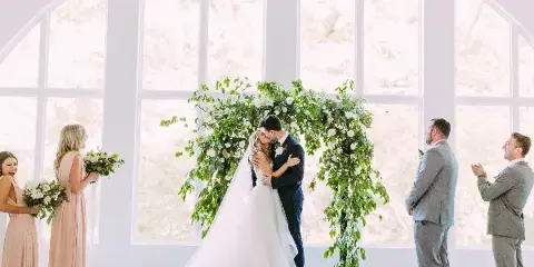 A bride and groom share a kiss at the altar during a wedding ceremony, surrounded by bridesmaids and groomsmen.