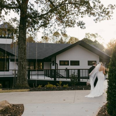 A bride and groom standing on a patio in front of a white venue building with a dark roof.