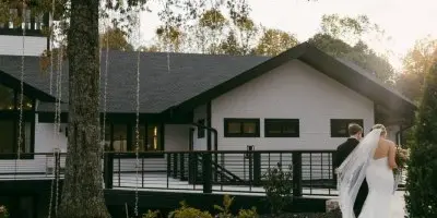 A bride and groom standing on a patio in front of a white venue building with a dark roof.