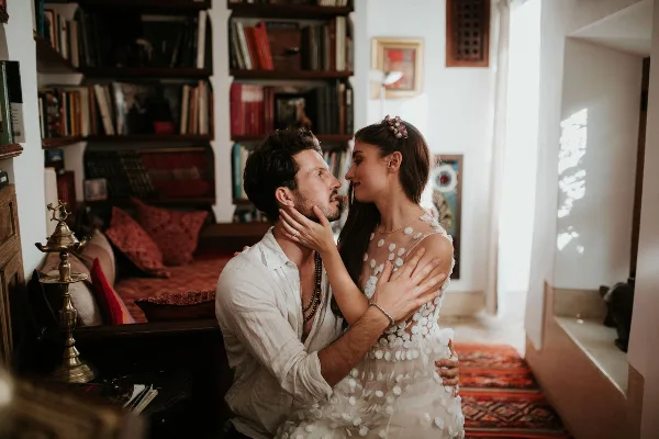 A bride and groom sharing an intimate moment in a room with bookshelves and warm lighting.