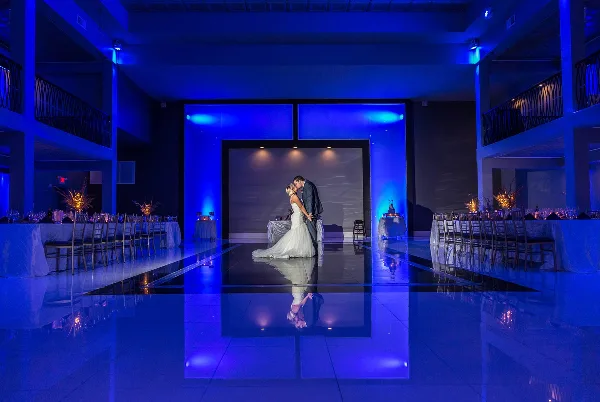 A bride and groom dancing on a reflective floor in a large banquet hall with blue ambient lighting.