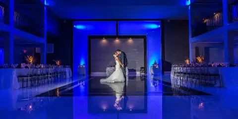 A bride and groom dancing on a reflective floor in a large banquet hall with blue ambient lighting.