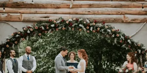 A bride and groom exchange vows under a floral arch during their wedding ceremony, surrounded by guests.