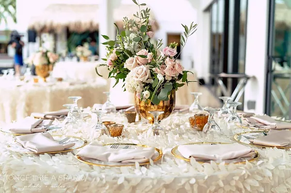 Elegant wedding reception table setting featuring a floral centerpiece in a gold vase, white rose petals, and fine dining ware.
