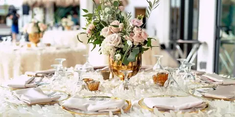 Elegant wedding reception table setting featuring a floral centerpiece in a gold vase, white rose petals, and fine dining ware.