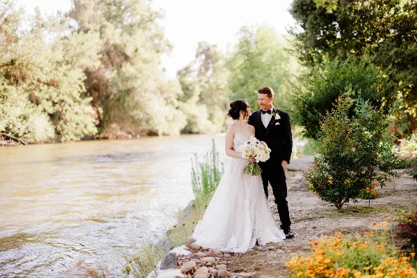 Bride and groom standing together on a grassy riverbank with a flowing river and trees in the background.