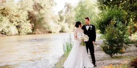 Bride and groom standing together on a grassy riverbank with a flowing river and trees in the background.