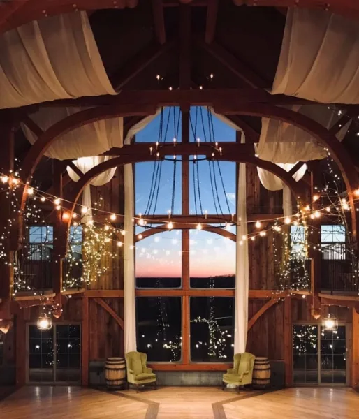 Interior view of a rustic barn venue featuring large arched windows, wooden beams, and string lights.