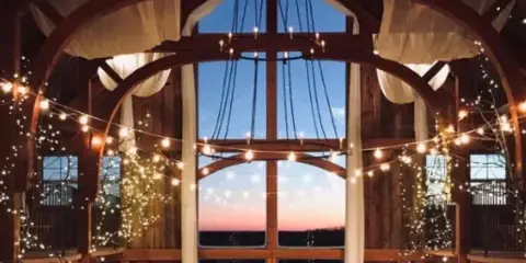Interior view of a rustic barn venue featuring large arched windows, wooden beams, and string lights.