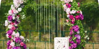 A floral wedding arch decorated with pink and purple roses and greenery, set outdoors on a lawn.