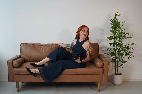 A woman with red hair and tattoos sits on a brown leather couch holding a professional video camera.