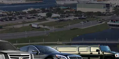 A fleet of luxury vehicles, including a black Cadillac SUV, a black Mercedes-Benz sedan, and a black stretch limousine, parked in front of a cruise ship port.