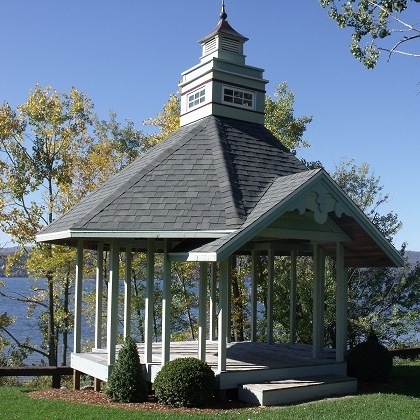 A white gazebo with a shingled roof and cupola stands on a lawn overlooking a lake.