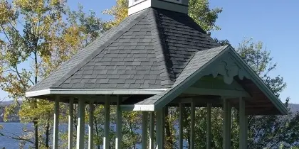 A white gazebo with a shingled roof and cupola stands on a lawn overlooking a lake.