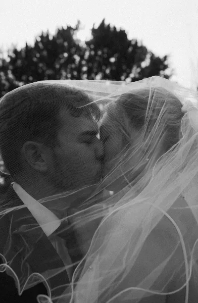 A black and white, cinematic photograph of a bride and groom sharing a kiss, with the bride's veil draped over both of them.