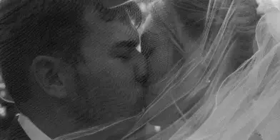 A black and white, cinematic photograph of a bride and groom sharing a kiss, with the bride's veil draped over both of them.