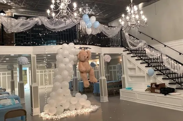 Interior of an event venue featuring a staircase, chandeliers, and a large balloon arch with a teddy bear.