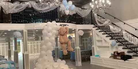 Interior of an event venue featuring a staircase, chandeliers, and a large balloon arch with a teddy bear.