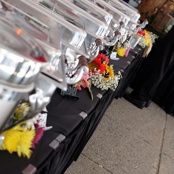 A catering buffet table featuring silver chafing dishes and colorful floral decorations.
