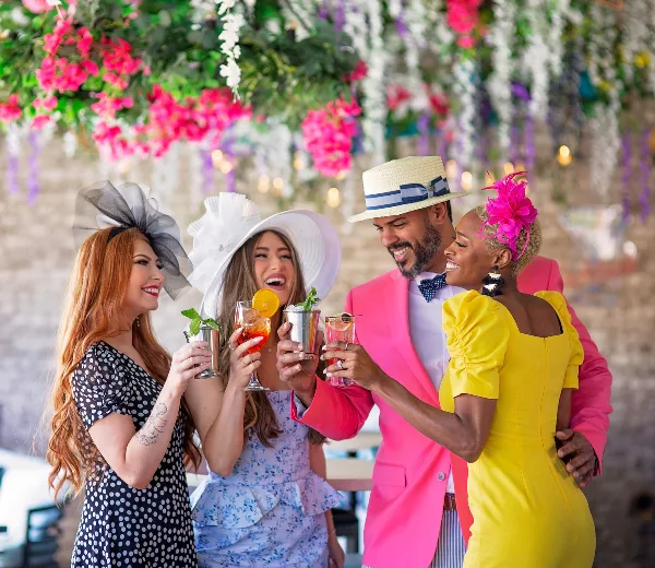 A group of four people in colorful formal wear toasting with drinks in front of a floral backdrop.