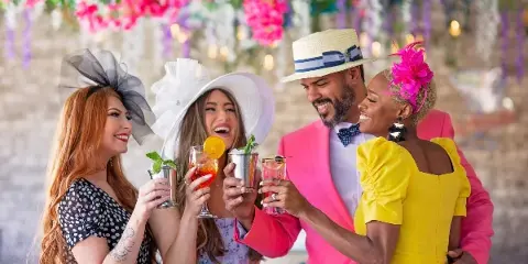 A group of four people in colorful formal wear toasting with drinks in front of a floral backdrop.