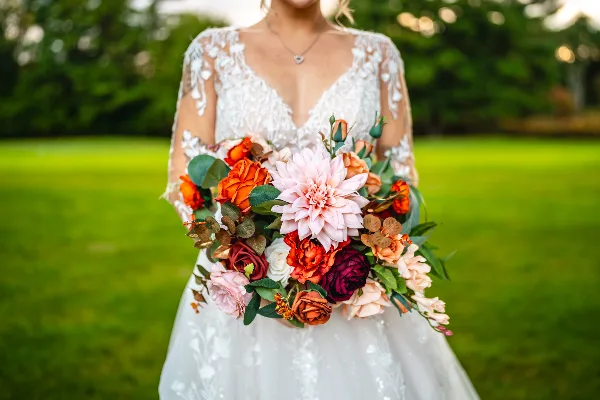 A bride in a white lace dress holding a colorful bouquet of flowers in a grassy outdoor setting.