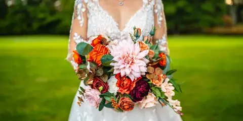 A bride in a white lace dress holding a colorful bouquet of flowers in a grassy outdoor setting.