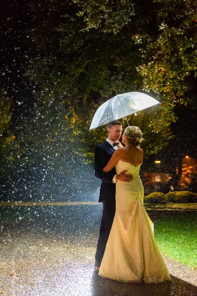 A bride and groom embracing under a clear umbrella in the rain at night, illuminated by soft light.