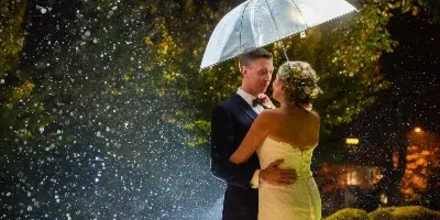 A bride and groom embracing under a clear umbrella in the rain at night, illuminated by soft light.