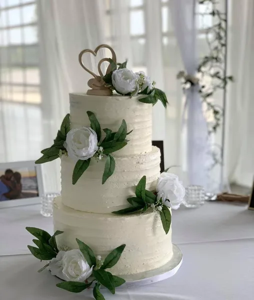 A three-tiered white wedding cake with textured frosting, decorated with white peonies, greenery, and a wooden heart topper.