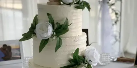 A three-tiered white wedding cake with textured frosting, decorated with white peonies, greenery, and a wooden heart topper.