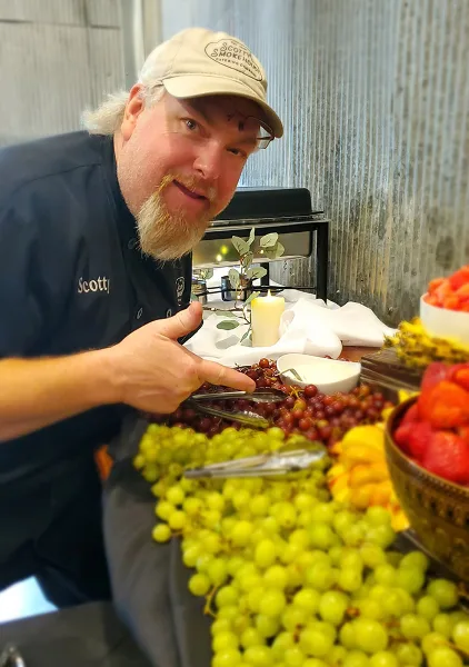 A man wearing a Scotty's Smokehouse Catering cap and shirt stands behind a catering table filled with fresh fruit and serving tongs.