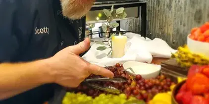 A man wearing a Scotty's Smokehouse Catering cap and shirt stands behind a catering table filled with fresh fruit and serving tongs.