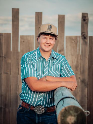 A professional portrait of a young man wearing a cap and a patterned shirt, leaning against a wooden fence.
