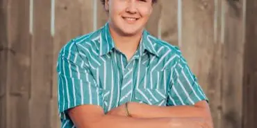 A professional portrait of a young man wearing a cap and a patterned shirt, leaning against a wooden fence.
