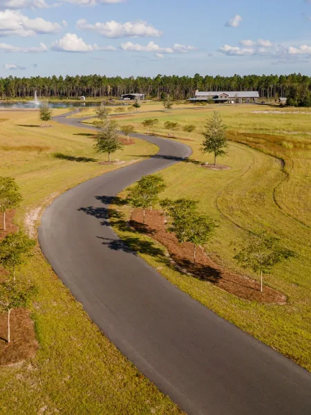 Aerial view of a rural plantation venue with a winding paved road, open grassy fields, a lake with a fountain, and a lodge building in the distance.