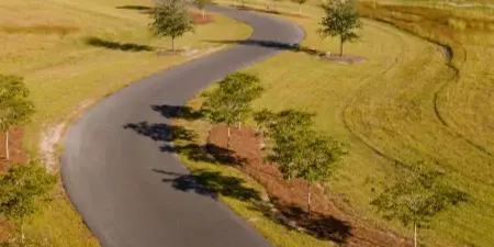 Aerial view of a rural plantation venue with a winding paved road, open grassy fields, a lake with a fountain, and a lodge building in the distance.