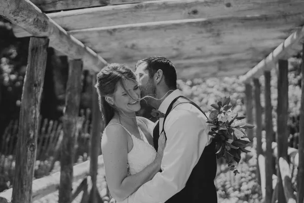 Black and white photograph of a bride and groom embracing under a wooden pergola structure.